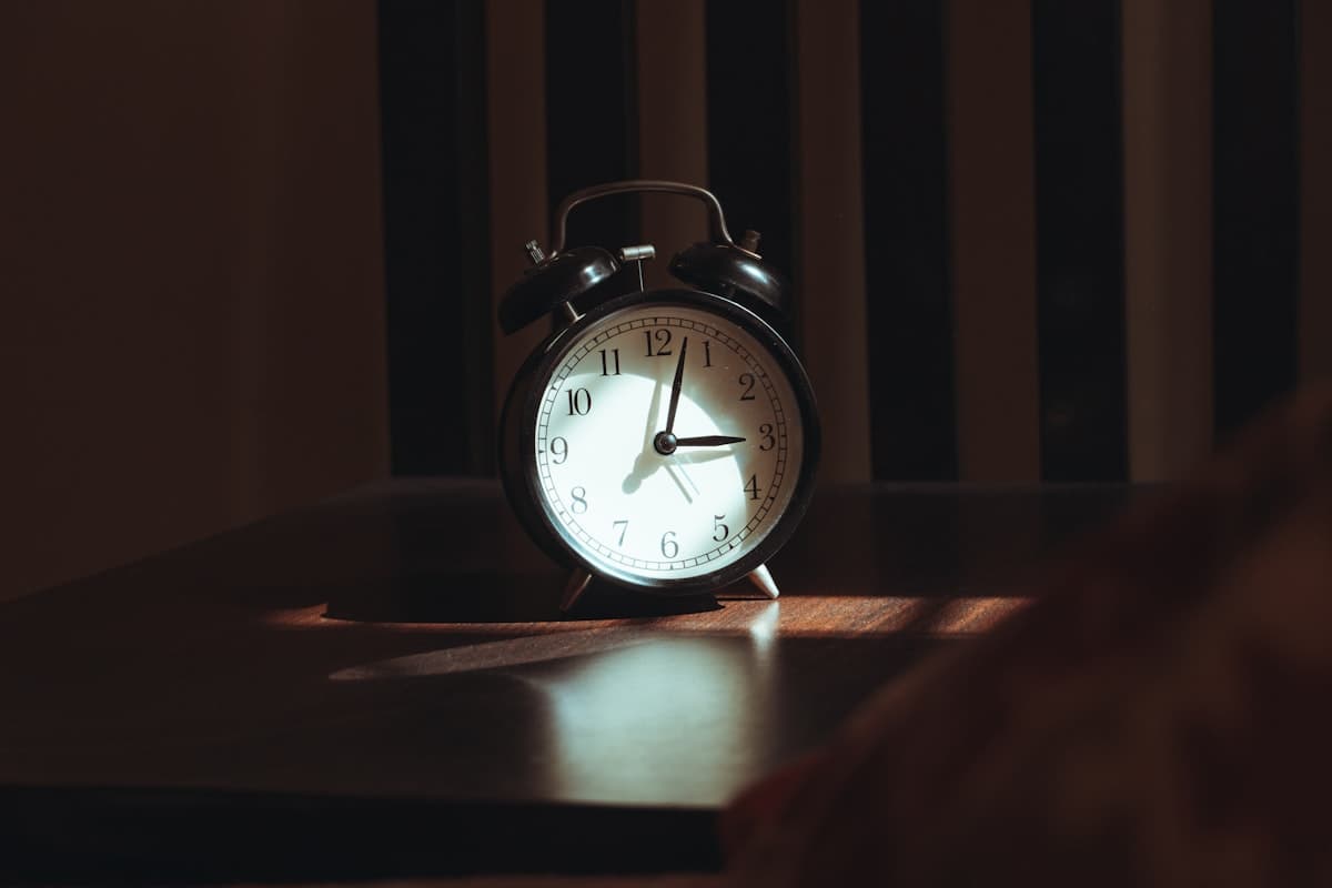 An alarm clock reading past 3am, lit by striped light through blinds.
