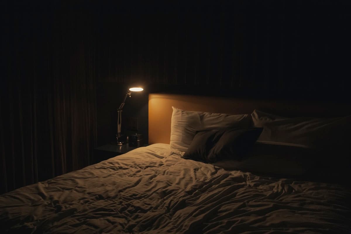 A single desk lamp glowing beside a bed in an otherwise dark bedroom.